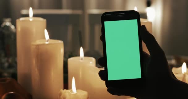 Male Hand is Holding Smartphone with Green Screen and tapping. Lighted candles in the background — Stock Video