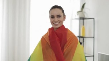 Portre genç kadın evde ayakta Lgbt bayrağı giyiyor. Kameraya bakıyor, gülümsüyor..