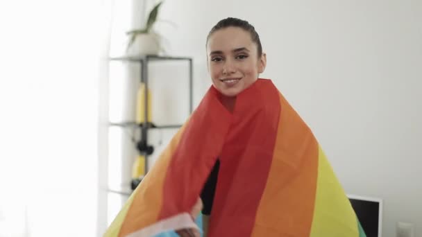 Portret jonge vrouw dragen LGBT vlag staande thuis. Ze kijkt naar de camera, glimlachend. — Stockvideo