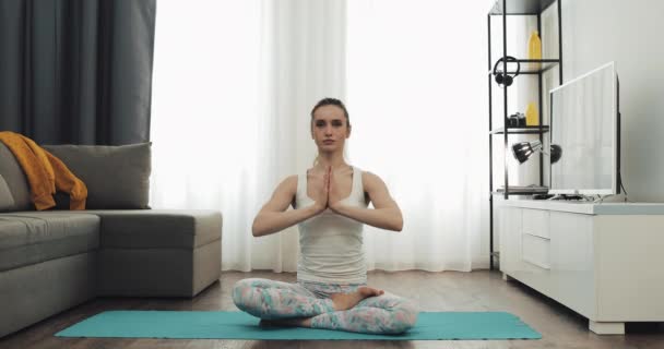 Portrait d'une jeune femme pratiquant le yoga assise sur un tapis en pose de lotus à la maison . — Video