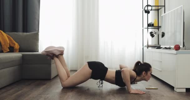 Eine junge Fitness-Frau in sportlicher Trainingskleidung macht Liegestützübungen, während sie eine Stoppuhr auf ihrem Smartphone bedient. Sie trainiert zu Hause. gesunder und sportlicher Lebensstil. — Stockvideo