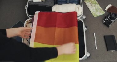 Womans hands packing suitcase for a journey on the bed at home. The girl puts the flag of the LGBT in a suitcase. Travel preparations.