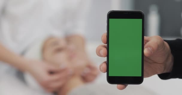 Close-up de um Mans Hand Holding Green Screen Smartphone no fundo de procedimentos de osmetologia. O Conceito de Beleza, Medicina e Cuidado do Rosto — Vídeo de Stock