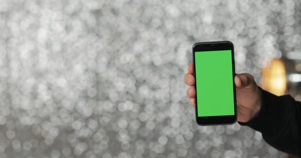 Nahaufnahme eines Mannes, der ein grünes Bildschirm-Smartphone auf dem silbernen Pailletten-Hintergrund hält — Stockvideo