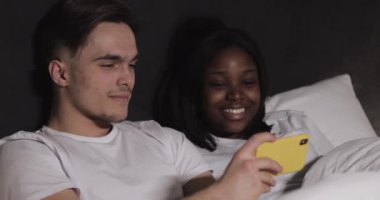 Mutlu çok ırklı çift gece yatakta yatan sarı akıllı telefon birlikte video izliyor. Gülüyorlar ve eğleniyorlar. İlişkiler, aile, sosyal ağ, iyi bir eğlence konsepti.