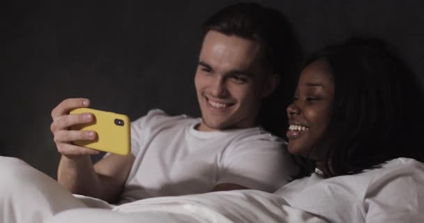 Gelukkig multi-etnisch paar kijken samen video op de gele smartphone liggend in bed 's nachts. Relaties, familie, sociaal netwerk, een goed tijdverdrijf concept. — Stockvideo