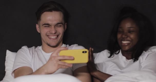 夜にベッドに横たわっている黄色いスマートフォンで一緒にビデオを見て幸せな多民族のカップル。彼らは笑って楽しんでいます。関係、家族、ソーシャルネットワーク、良い娯楽の概念. — ストック動画