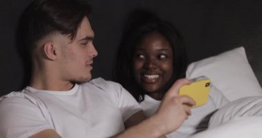 Mutlu çok ırklı çift gece yatakta yatan sarı akıllı telefon birlikte video izliyor. Gülüyorlar ve eğleniyorlar. İlişkiler, aile, sosyal ağ, iyi bir eğlence konsepti.