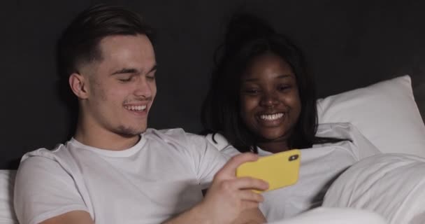 Happy multi-ethnic couple watching video together on the yellow smartphone lying in bed at night. They laughing and having fun. Relationships, family, social network, a good pastime concept. — Stock Video