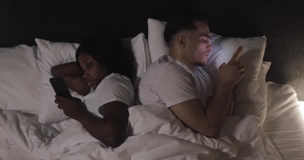 結婚した若い多民族カップルは、夜にベッドに横たわって、スマートフォンで入力したりスクロールしたりします。屋内。トップビュー. — ストック動画