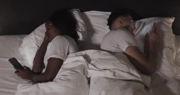 Getrouwd jong multi-etnisch echtpaar liggend in het bed terug naar achteren 's nachts en typen of scrollen op smartphones. Indoor. Top View. — Stockvideo