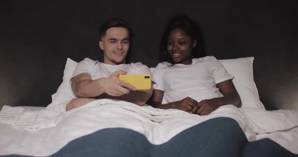 Gelukkig multi-etnisch paar kijken samen video op de gele smartphone liggend in bed 's nachts. Ze lachen en plezier hebben. Relaties, familie, sociaal netwerk, een goed tijdverdrijf concept. — Stockvideo