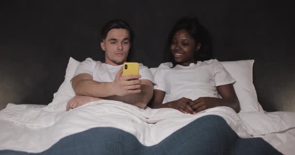 Ευτυχισμένο πολυεθνικό ζευγάρι βλέποντας βίντεο μαζί στο κίτρινο smartphone που βρίσκεται στο κρεβάτι τη νύχτα. Γελάνε και διασκεδάζουν. Σχέσεις, οικογένεια, κοινωνικό δίκτυο, μια καλή ιδέα για χόμπι. — Αρχείο Βίντεο