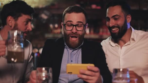 Three old friends watching video on smartphone and drinking draft beer in the sport bar. Friends laughing and discuss the news in beer pub. — Stock Video