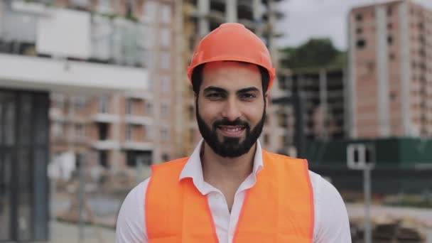 Bina yerinde inşaat işçisi portre fotoğraf makinesi gülümsüyor. İnşaatçı modern bir binanın arka plana karşı duruyor. Yavaş hareket. — Stok video