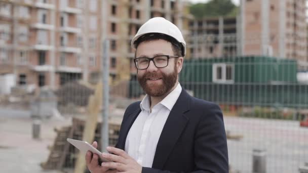 Retrato de jovem empresário com tablet no canteiro de obras sorrindo olhando para a câmera vestindo um capacete de segurança e terno. Engenheiro, arquiteto, construtor, empresário . — Vídeo de Stock