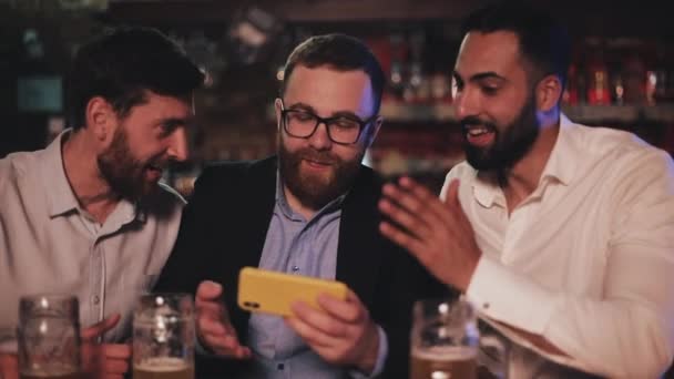 Três velhos amigos assistindo vídeo no smartphone e bebendo cerveja no bar esportivo. Amigos rindo e discutir as notícias no pub de cerveja . — Vídeo de Stock