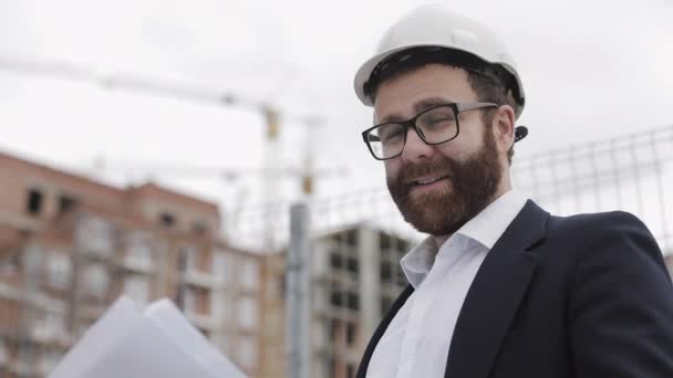 Portrait of handsome young engineer on building site looking at the camera. The builder with construction project stands against the backdrop of a modern building. Slow motion. — Stock Video