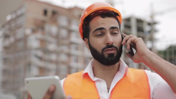 Інженер будівельника інспектор розмовляє на смартфоні і працює на планшеті, стоячи на фоні будівельного майданчика. Концепція професії . — стокове відео