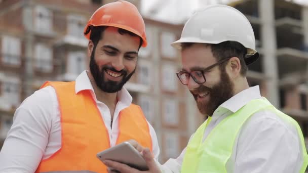 Smiling constructor y arquitecto hombre están discutiendo el plan de construcción del moderno centro de negocios de pie cerca de la obra. Comprueban el trabajo con una tableta . — Vídeos de Stock