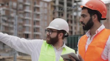 İnşaatçı ve mimar adam inşaat alanı yakınında modern iş merkezi ayakta inşaat planını tartışıyorlar. İşleri bir tabletle kontrol ediyorlar.