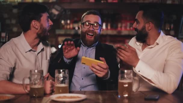 三位老朋友在智能手机上观看视频, 并在体育酒吧喝生啤酒。朋友们在啤酒酒吧里笑着讨论这个消息. — 图库视频影像
