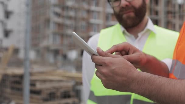 Close up de mãos de arquiteto usando tablet perto do canteiro de obras. O construtor e arquiteto homem estão discutindo o plano de construção do moderno centro de negócios . — Vídeo de Stock