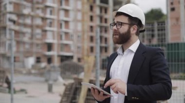 Mimar adam şantiye ve analiz şeması proje planı üzerinde tablet ile ayakta iş elbisesi giyiyor. Dış, yavaş çekim.
