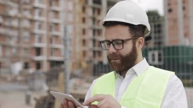 Beyaz kasklı, tabletle çalışan ve kameraya bakan başarılı genç bir iş adamının portresi. İnşaatçı modern bir binanın arka planında duruyor..