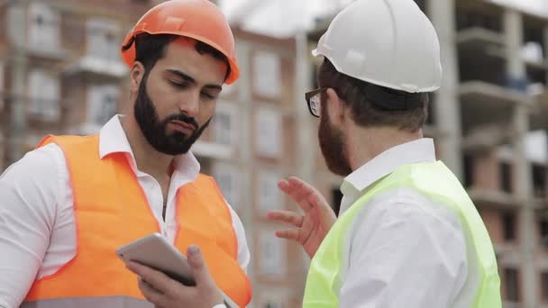 İnşaatçı ve mimar adam inşaat alanı yakınında modern iş merkezi ayakta inşaat planını tartışıyorlar. İşleri bir tabletle kontrol ediyorlar. — Stok video