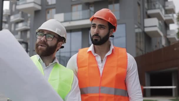 O construtor e arquiteto homem estão discutindo o plano de construção do moderno centro de negócios andando perto do canteiro de obras. Eles verificam o trabalho com a elaboração arquitetônica . — Vídeo de Stock