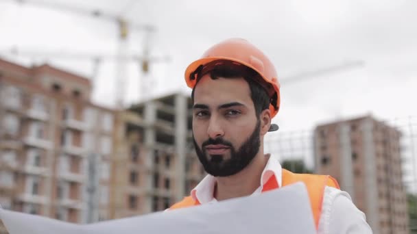 Retrato de trabalhador da construção sorridente em capacete laranja olhando para a câmera. O construtor com projeto de construção fica no fundo do canteiro de obras. Movimento lento . — Vídeo de Stock