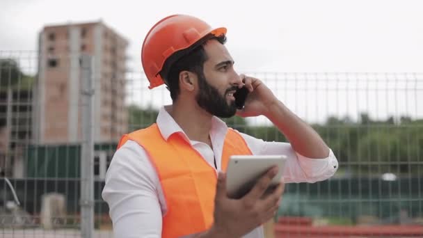 Инспектор строительства инженер разговаривает по смартфону и работает на планшете стоя на фоне строительной площадки. Концепция профессии . — стоковое видео