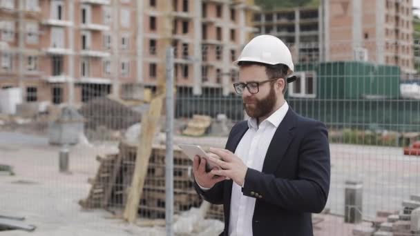 Portrait of young businessman with tablet on construction site smiling looking to camera wearing a safety helmet and suit. Engineer, architect, builder, businessman. — Stock Video