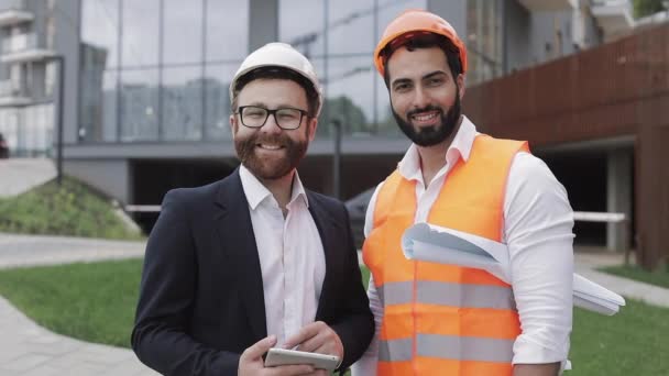 Porträtt av Happy Builder och affärsman tittar på kameran står mot bakgrund av en modern byggnad. Yrken, byggande, arbetstagare, arkitekt koncept. — Stockvideo