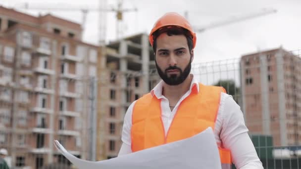 Ritratto di muratore in casco arancione che guarda la macchina fotografica. Il costruttore con progetto di costruzione si trova sullo sfondo del cantiere. Rallentatore . — Video Stock