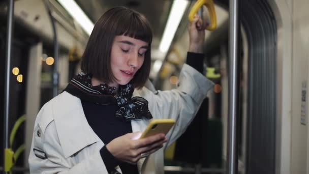 Portrait de jeune femme hipster tient la main courante, en utilisant le smartphone debout dans les transports publics. Fond de lumières de ville .