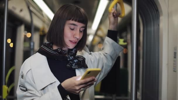 Retrato de mujer hipster joven sostiene la barandilla, utilizando el teléfono inteligente de pie en el transporte público. Ciudad luces fondo . — Vídeo de stock
