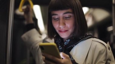 Genç hipster kadın portresi, toplu taşıma ayakta akıllı telefon kullanarak, korkuluk tutar. Şehir ışıkları arka plan.