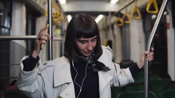 Porträt einer jungen Frau im Mantel mit Kopfhörern, die Musik hört und lustig in öffentlichen Verkehrsmitteln tanzt. er hält den Handlauf. — Stockvideo