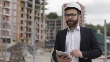 Mimar adam şantiye ve analiz şeması proje planı üzerinde tablet ile yürüyüş iş elbisesi giyiyor. Dış, yavaş çekim.