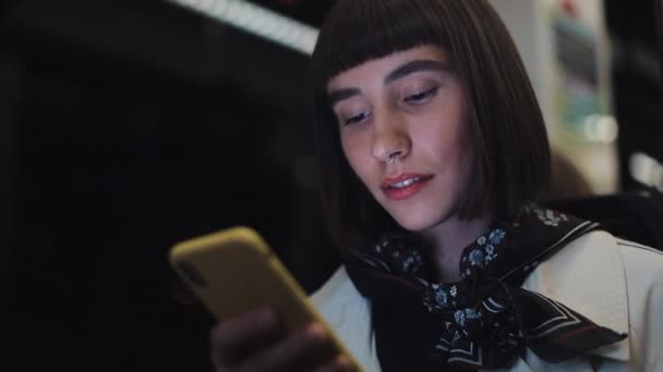 Junge Hipsterfrau mit Smartphone in öffentlichen Verkehrsmitteln sitzend, Steadicam-Aufnahme. Zeitlupe. Hintergrund Stadtbeleuchtung. — Stockvideo