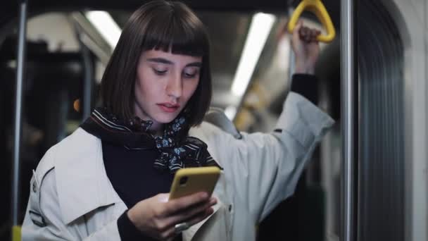 若いヒップスターの女性の肖像画は、公共交通機関に立ってスマートフォンを使用して、手すりを保持しています。都市ライトの背景. — ストック動画