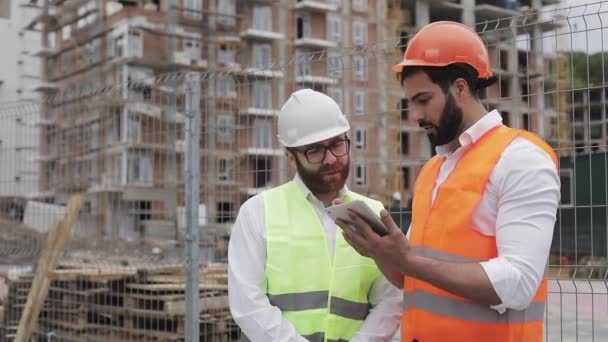 Budowniczy i architekt mężczyzna omawiają plan budowy nowoczesnego centrum biznesowego stojącego w pobliżu placu budowy. Sprawdzają pracę z tabletem. — Wideo stockowe