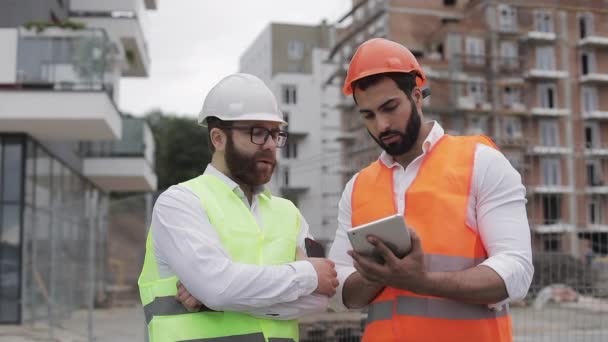 İnşaatçı ve mimar adam inşaat alanı yakınında modern iş merkezi ayakta inşaat planını tartışıyorlar. İşleri bir tabletle kontrol ediyorlar. — Stok video