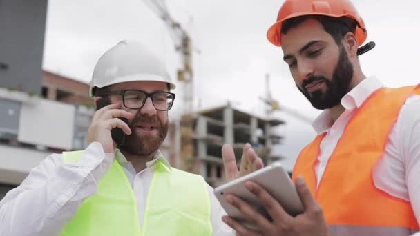 Happy Engineer spreekt op de mobiele telefoon op de bouwplaats en controleert het werk van de werknemer. Builder praat op smartphone. De bouwfase controleren — Stockvideo