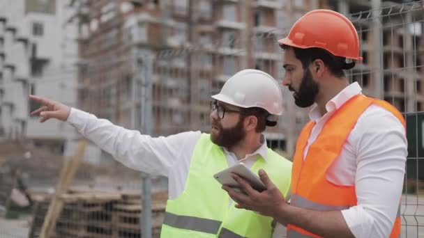 Budowniczy i architekt mężczyzna omawiają plan budowy nowoczesnego centrum biznesowego stojącego w pobliżu placu budowy. Sprawdzają pracę z tabletem. — Wideo stockowe