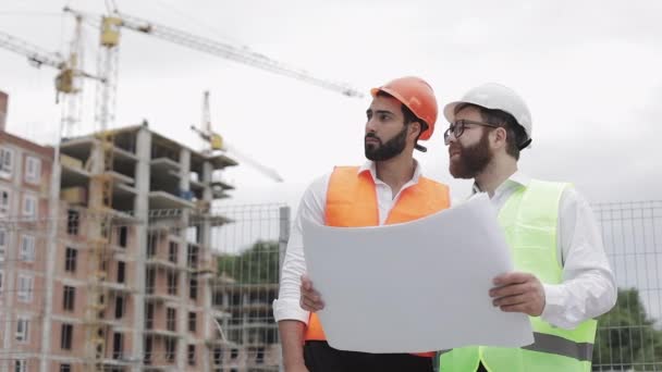 Yüksek katlı binanın şantiye veya şantiyemimar ile erkek inşaat mühendisi tartışma. Ellerinde inşaat çizimleri tutuyorlar.. — Stok video