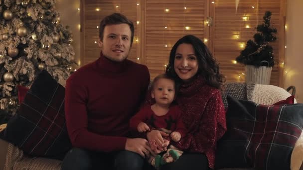 Portret szczęśliwej młodej rodziny siedzącej razem na kanapie w salonie w domu w dniu Bożego Narodzenia, uśmiechając się i patrząc na kamerę. Nowy rok i święta Bożego Narodzenia w koncepcji rodzinnej. — Wideo stockowe