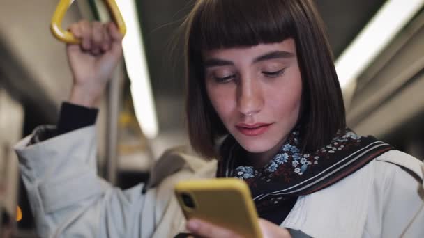 Retrato de la bella mujer sonriente en el transporte público sostiene la barandilla y la navegación en el teléfono inteligente amarillo. Fondo de luces de ciudad. Comunicación, carretera, concepto de viaje . — Vídeo de stock